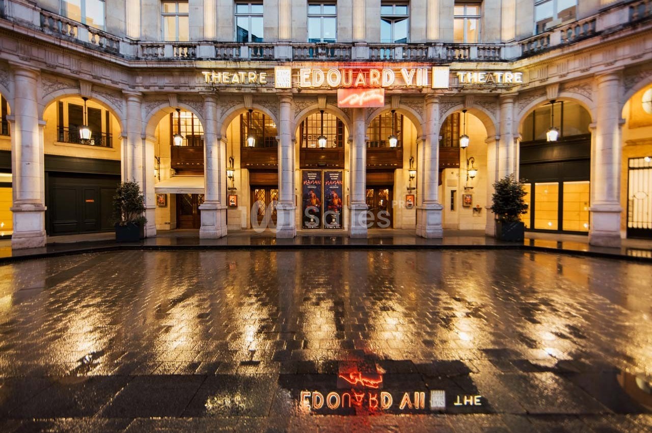 Façade illuminée du théâtre Édouard VII avec affiches et reflets sur le sol mouillé.
