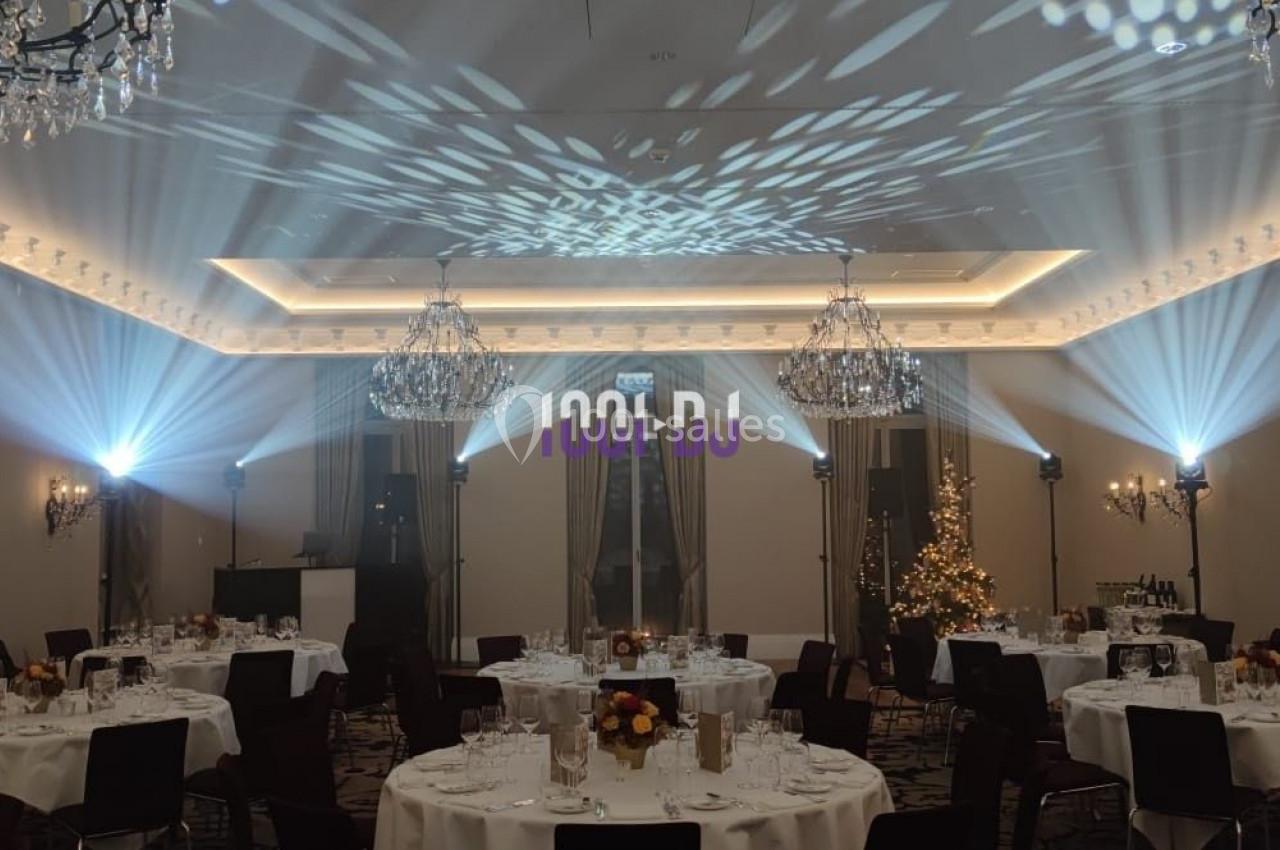 Salle de réception élégante avec tables rondes dressées, lustres et éclairage d'ambiance projeté au plafond.