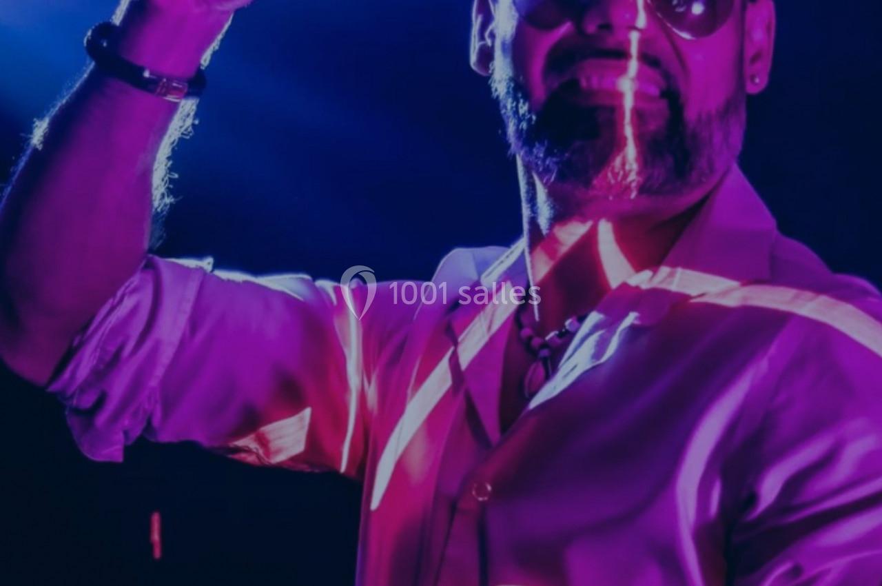 Un DJ en chemise blanche mixe sur scène sous des lumières violettes et roses, levant un doigt vers le public.