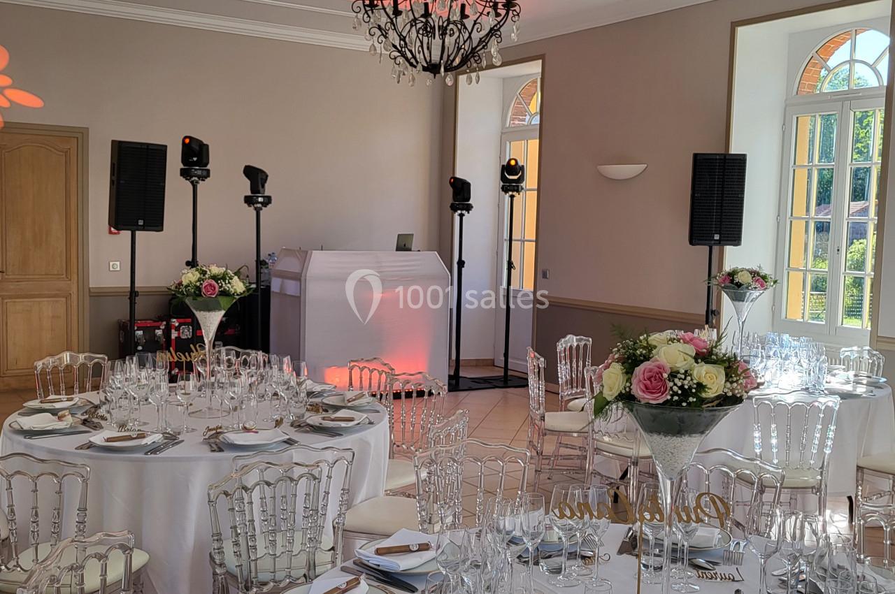 Salle de réception décorée avec des tables rondes dressées, des chaises transparentes et un espace pour la sonorisation.