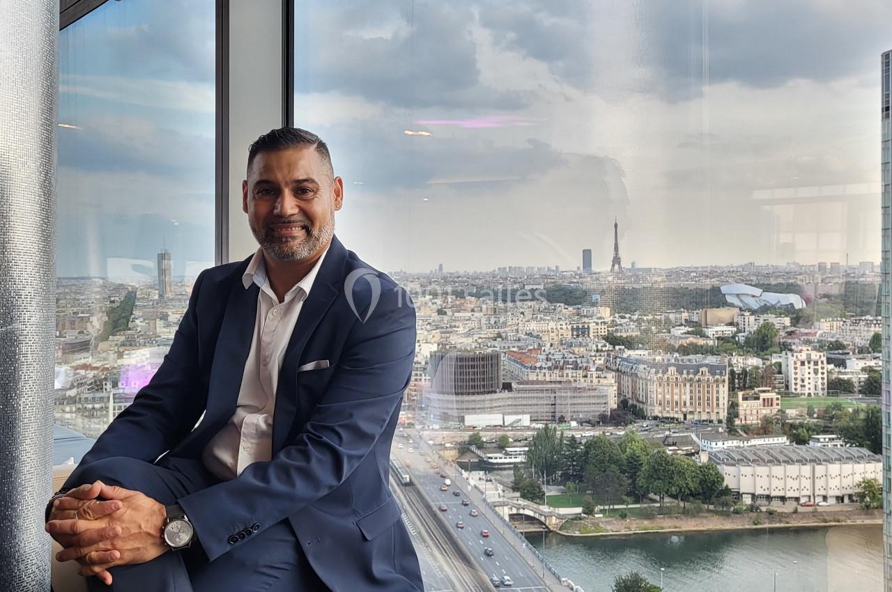 Homme en costume assis près d'une fenêtre avec vue panoramique sur une ville et la Tour Eiffel en arrière-plan.