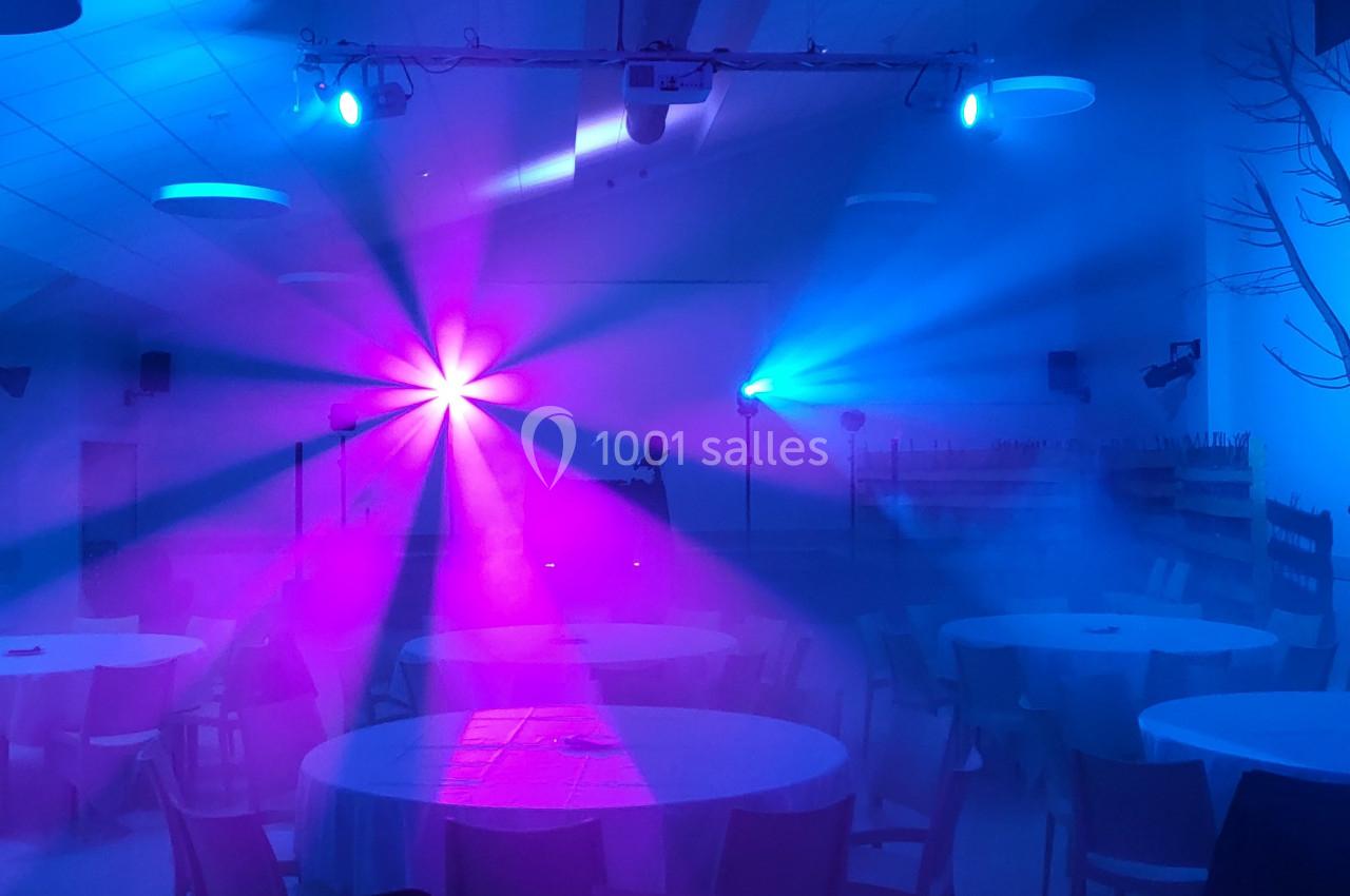 Salle décorée avec des tables rondes, éclairée par des lumières colorées bleues et roses créant une ambiance festive.