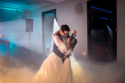 Un homme en chemise blanche et pantalon bleu anime un événement avec un micro dans une salle de réception.