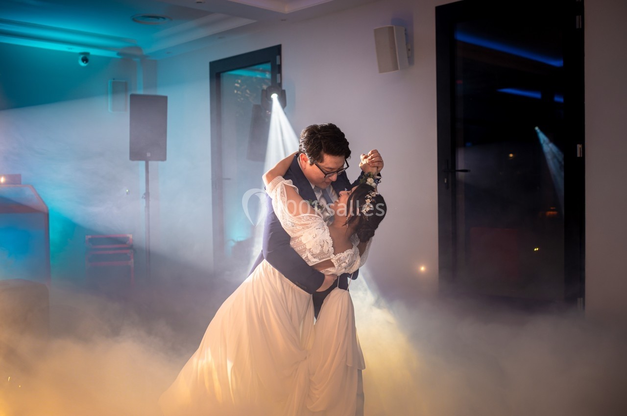 Un couple en tenue de mariage danse dans une salle éclairée par des projecteurs et entourée de fumée.
