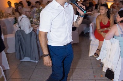 Un homme en chemise blanche et pantalon bleu anime un événement avec un micro dans une salle de réception.