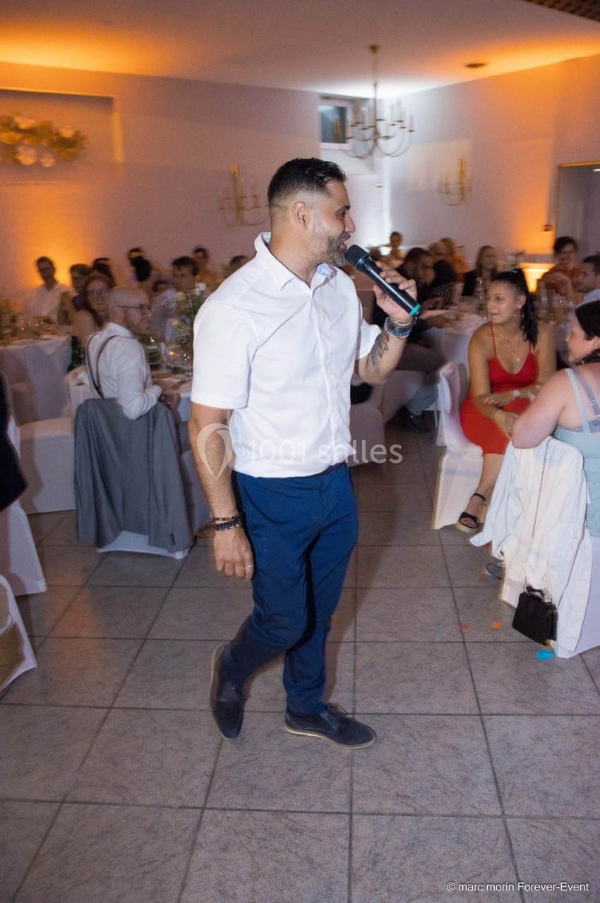 Un homme en chemise blanche et pantalon bleu anime un événement avec un micro dans une salle de réception.