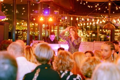 Un homme en chemise blanche et pantalon bleu anime un événement avec un micro dans une salle de réception.