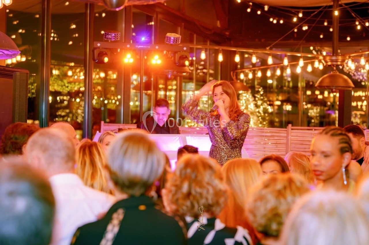 Une chanteuse en robe scintillante se produit devant un public dans une salle éclairée par des guirlandes lumineuses.