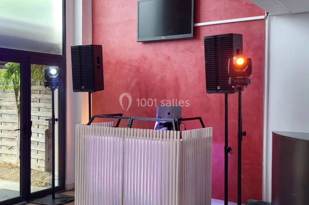 Poste de DJ avec une table lumineuse, deux enceintes sur pieds et des projecteurs dans une salle aux murs rouges.