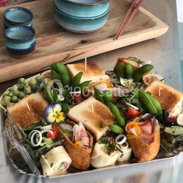 Plateau de bouchées variées comprenant sushis, légumes, fleurs comestibles et toasts, présenté sur une table avec vaisselle.