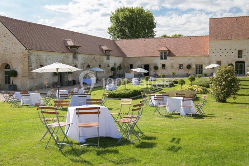 Cour d'une bâtisse en pierre avec tables et chaises installées sur une pelouse, entourée de parasols et de jardins.