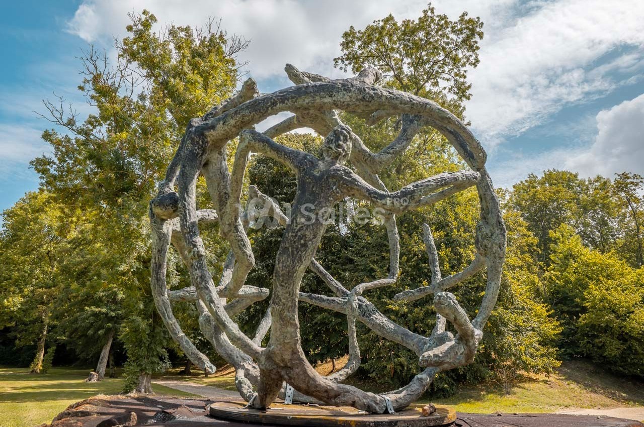 Sculpture en métal représentant des formes humaines entrelacées dans une sphère, exposée dans un parc verdoyant.