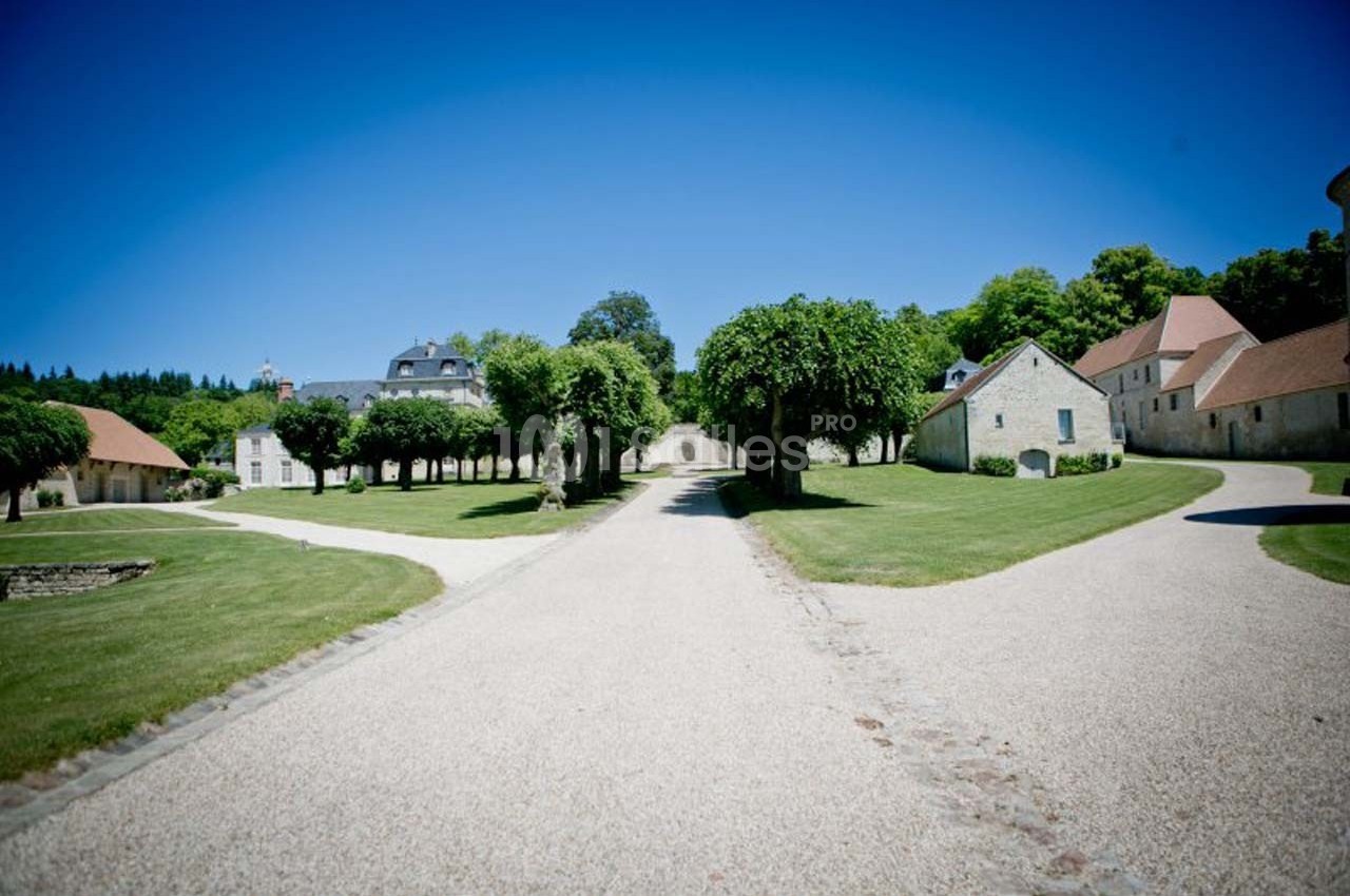 Allée bordée d'arbres menant à des bâtiments en pierre entourés de pelouses sous un ciel bleu dégagé.
