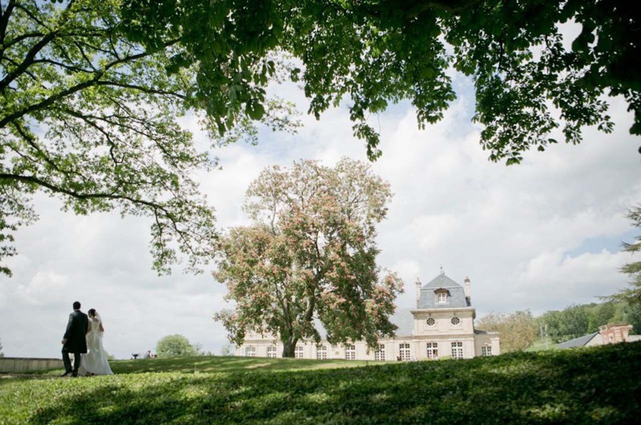 Un couple marche dans un parc verdoyant près d'un grand bâtiment historique sous un ciel partiellement nuageux.