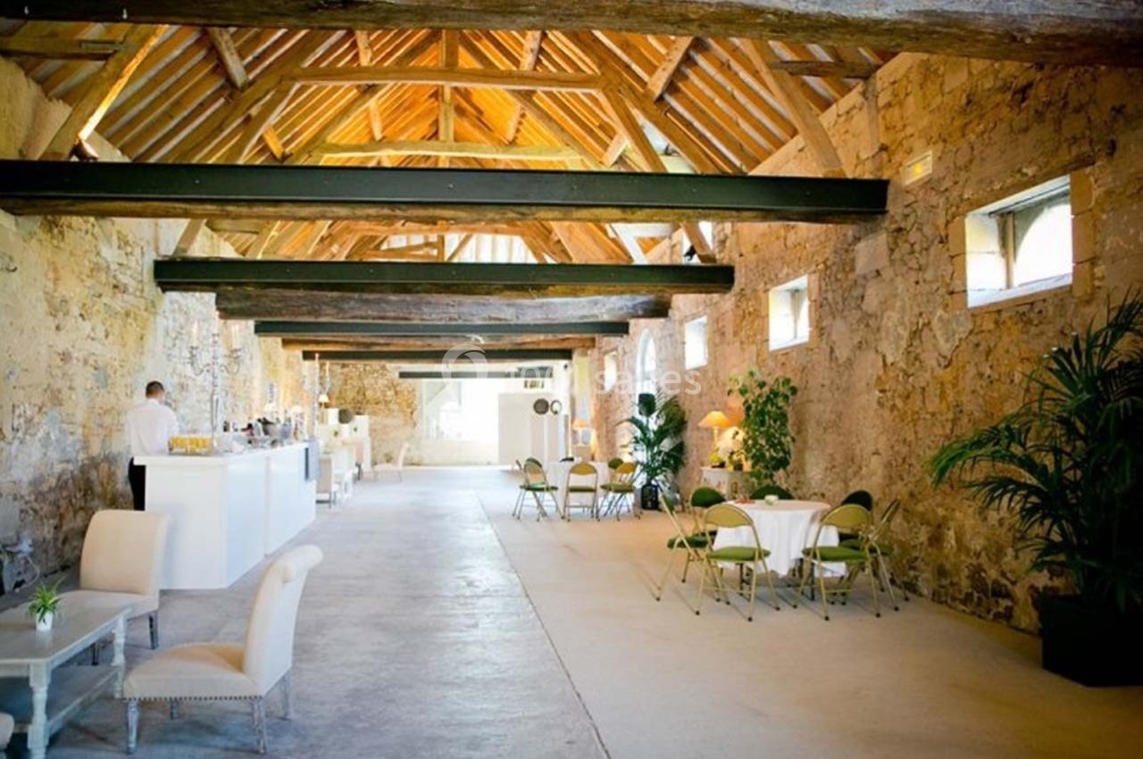 Salle en pierre avec poutres apparentes, aménagée avec un bar, des tables et des chaises, éclairée par la lumière naturelle.