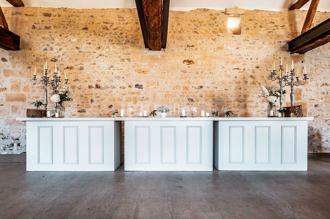 Trois comptoirs blancs décorés de bougies, chandeliers et fleurs, devant un mur en pierre dans une salle lumineuse.