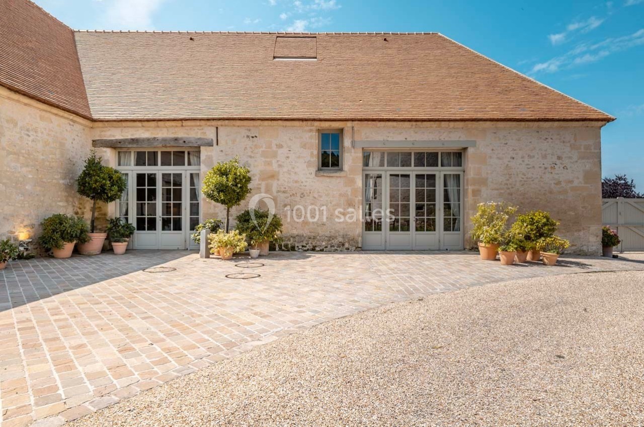 Façade en pierre d'un bâtiment avec toit en tuiles, grandes portes vitrées et plantes en pots sur une cour pavée.