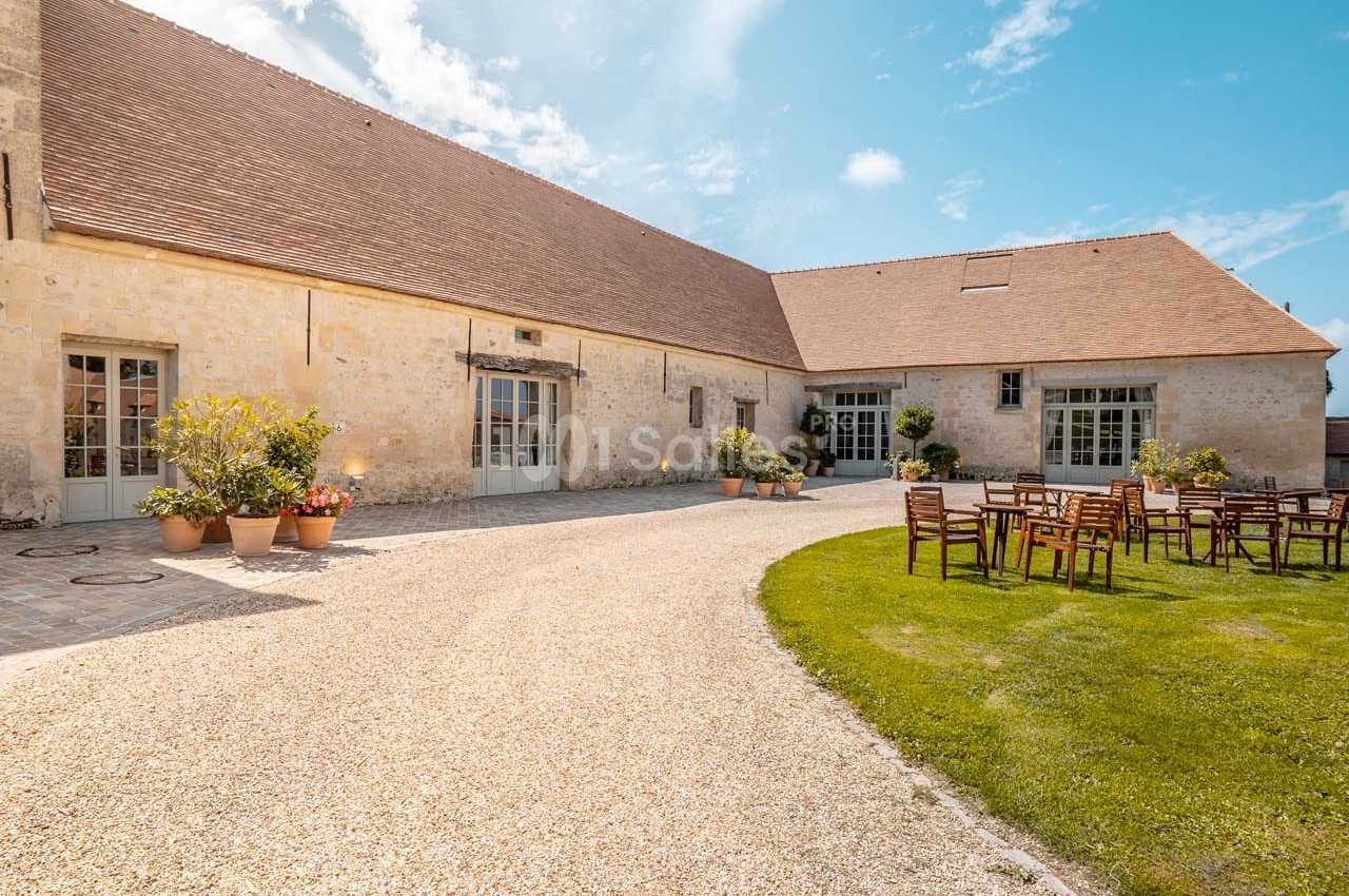 Cour extérieure d'un bâtiment en pierre avec des tables et chaises sur une pelouse, sous un ciel bleu.