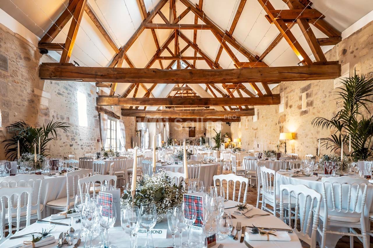 Salle de réception lumineuse avec poutres apparentes, tables décorées et chaises blanches disposées pour un événement.