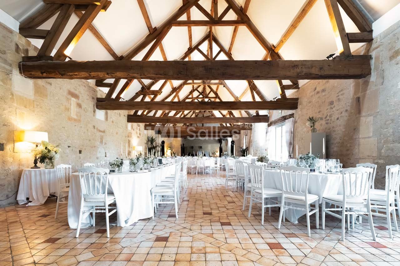 Salle de réception lumineuse avec poutres apparentes, tables dressées avec nappes blanches et chaises alignées.