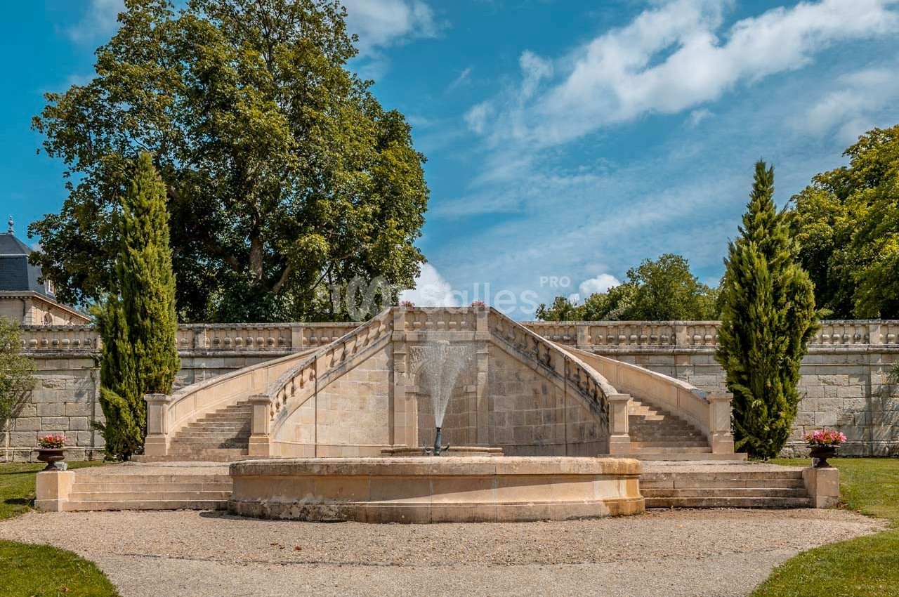Escalier en pierre symétrique avec fontaine centrale, entouré d'arbres et de verdure sous un ciel bleu.