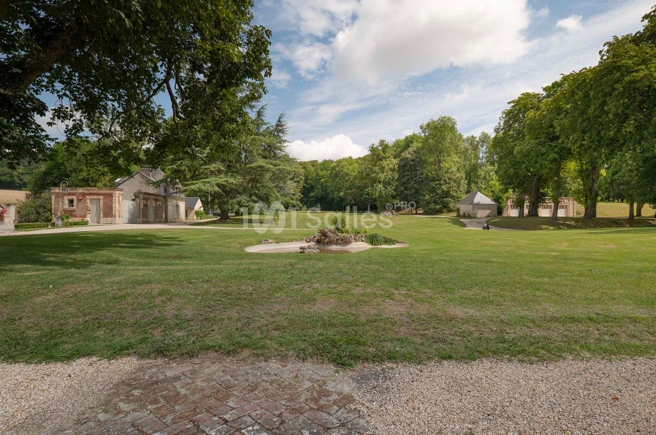 Grand jardin verdoyant avec pelouse, arbres, bâtiments en pierre et ciel partiellement nuageux en arrière-plan.