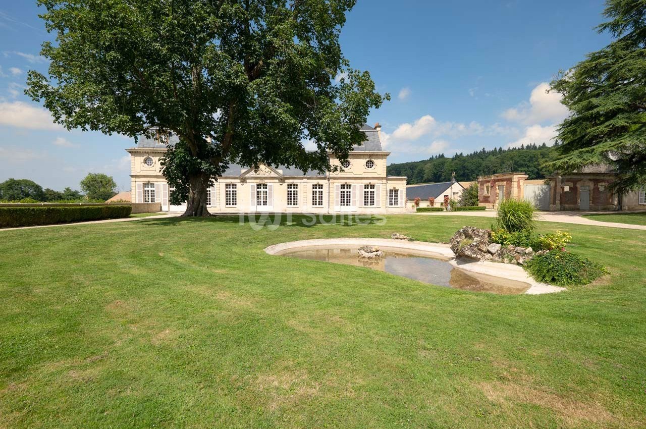 Grand bâtiment classique entouré d'une pelouse bien entretenue, avec un arbre imposant et un petit bassin au premier plan.