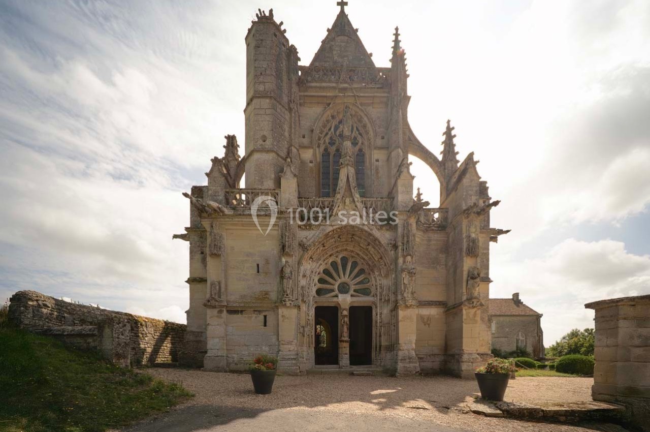 Façade d'une petite église gothique avec des détails sculptés, entourée de végétation et éclairée par une lumière douce.