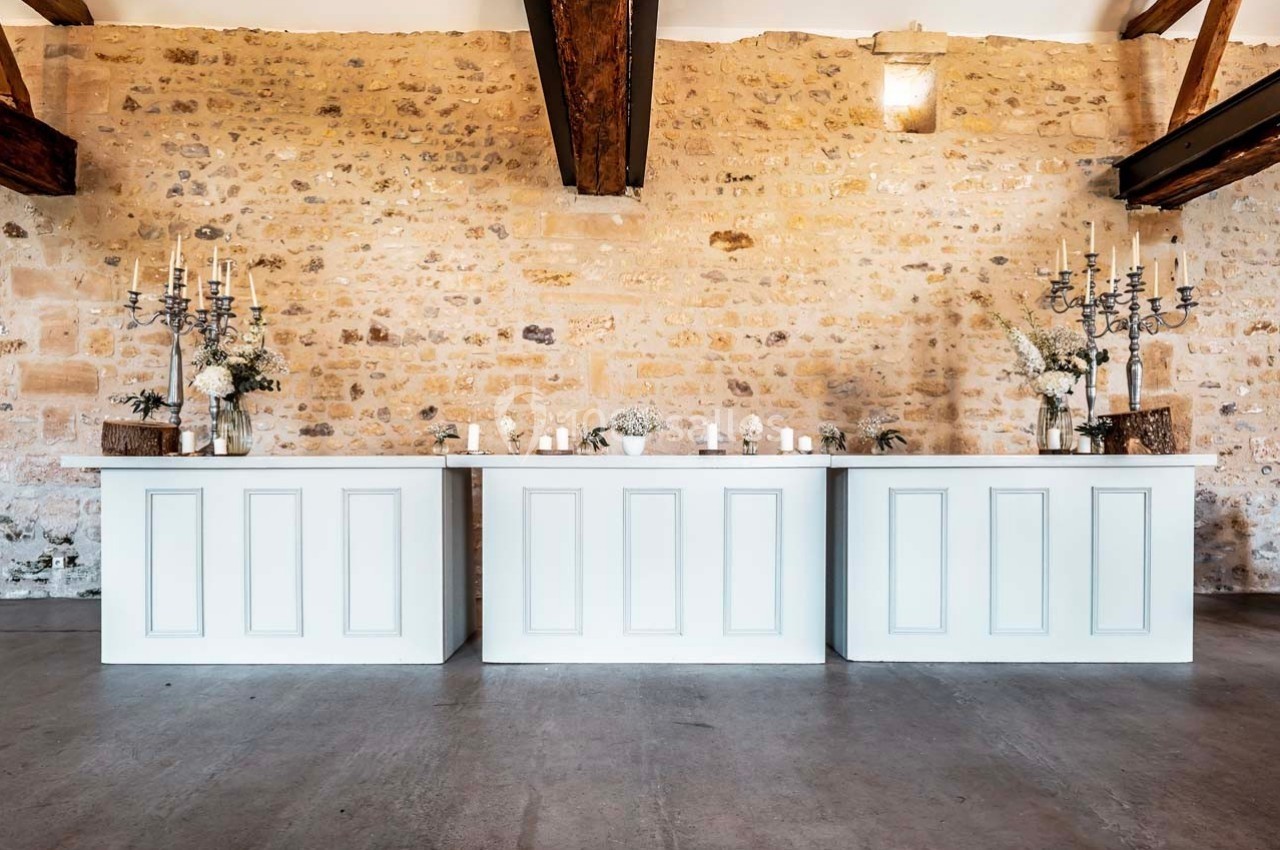 Trois comptoirs blancs alignés devant un mur en pierre, décorés de chandeliers et de fleurs.