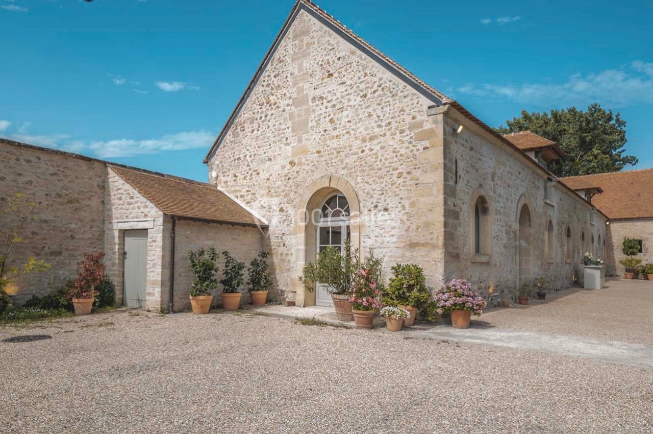 Bâtiment en pierre avec toit en tuiles, entouré de pots de fleurs, dans une cour en gravier sous un ciel bleu.