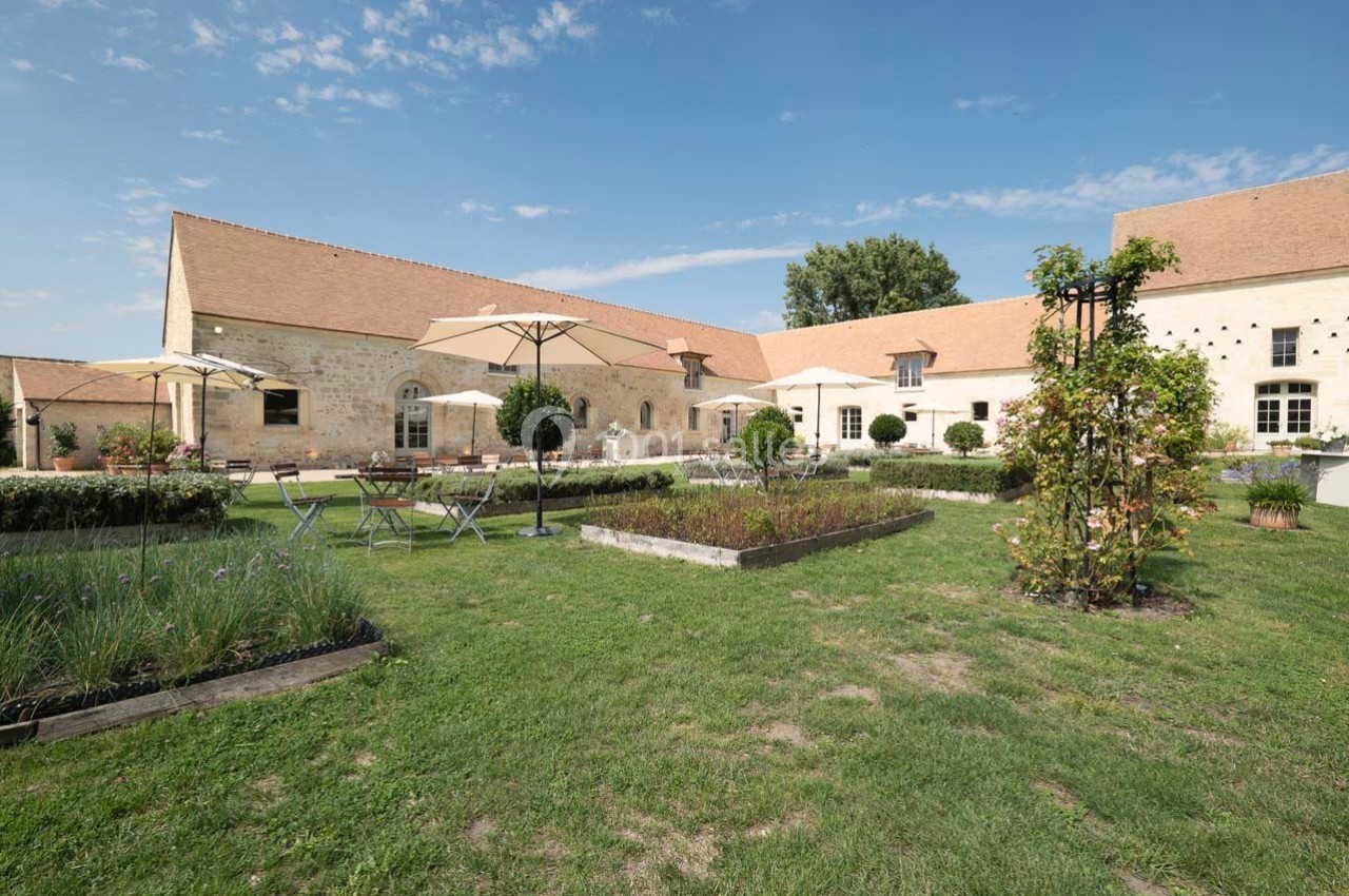 Cour d'un bâtiment en pierre avec jardin aménagé, tables, chaises et parasols sous un ciel dégagé.