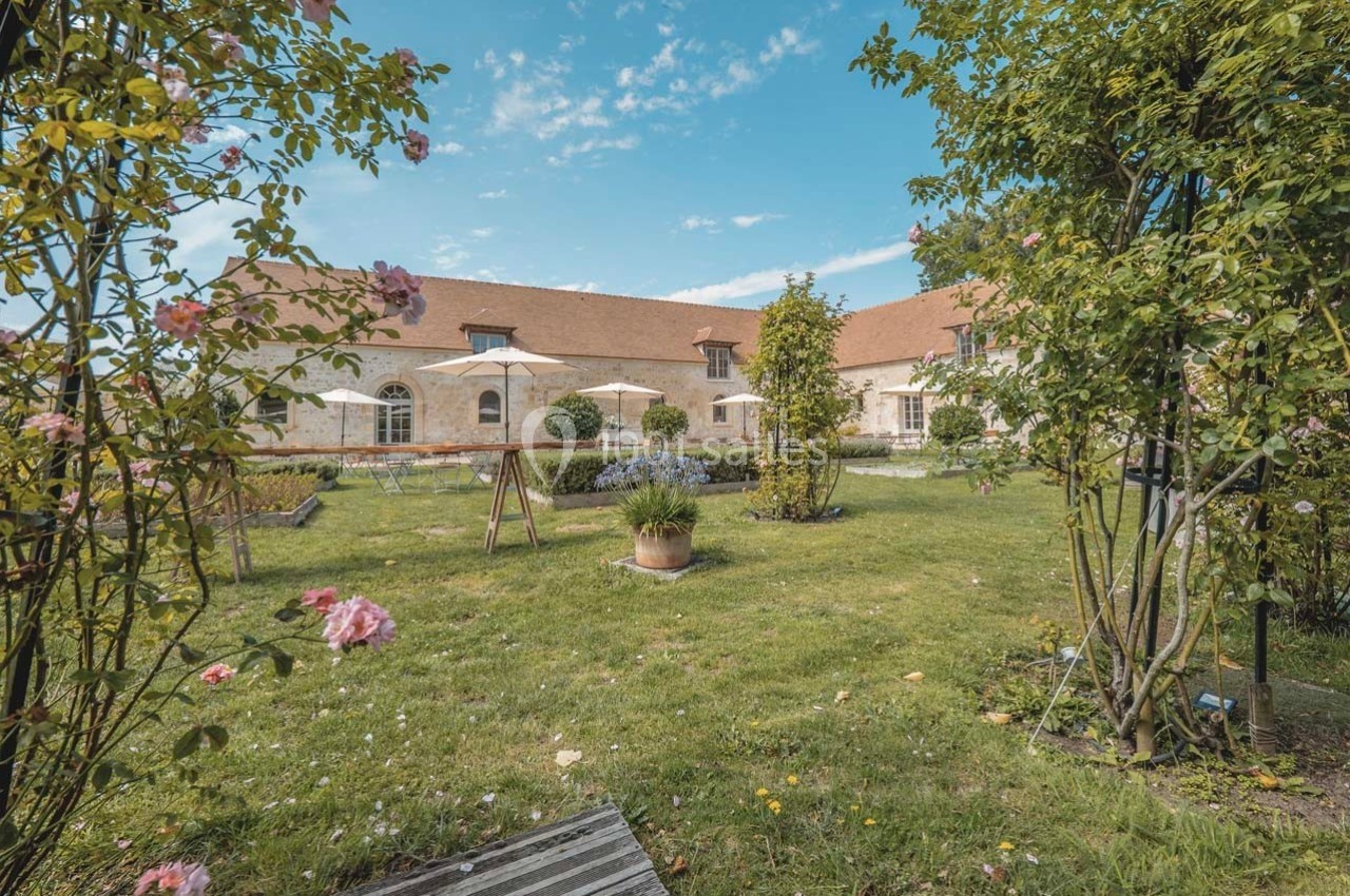 Jardin verdoyant avec pelouse, fleurs, arbres et terrasse aménagée devant un bâtiment en pierre sous un ciel bleu.