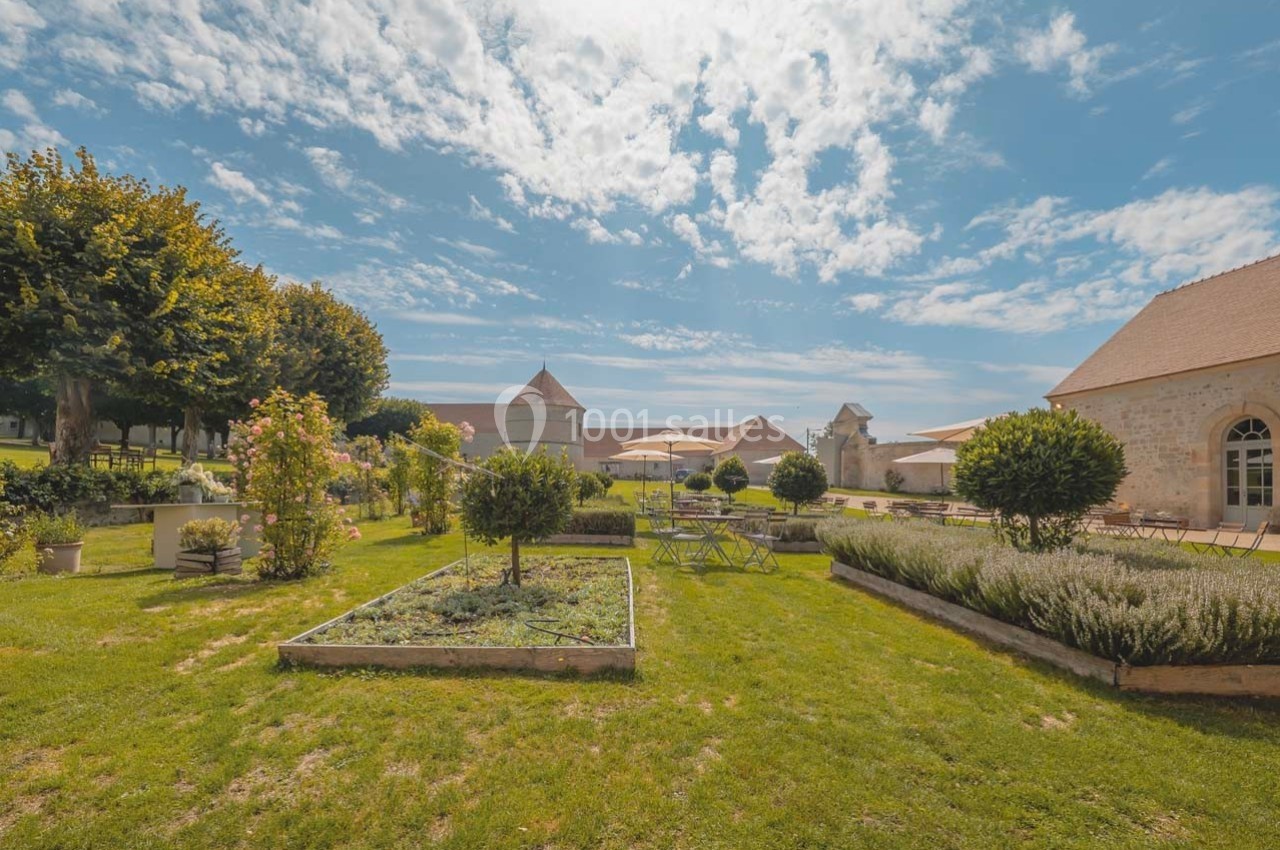 Jardin aménagé avec parterres de fleurs, pelouse et bâtiments en pierre sous un ciel bleu parsemé de nuages.