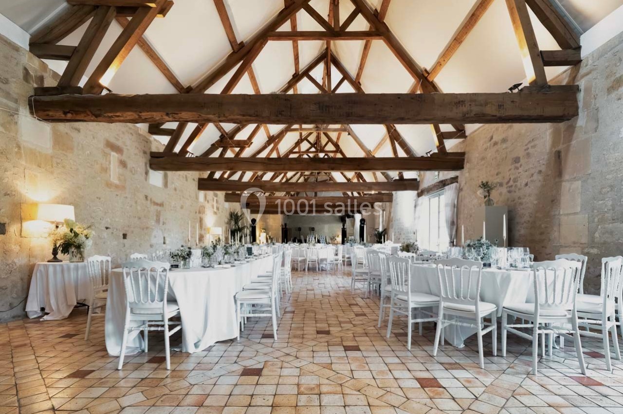 Salle de réception lumineuse avec poutres apparentes, tables dressées et chaises blanches sur un sol en carreaux.