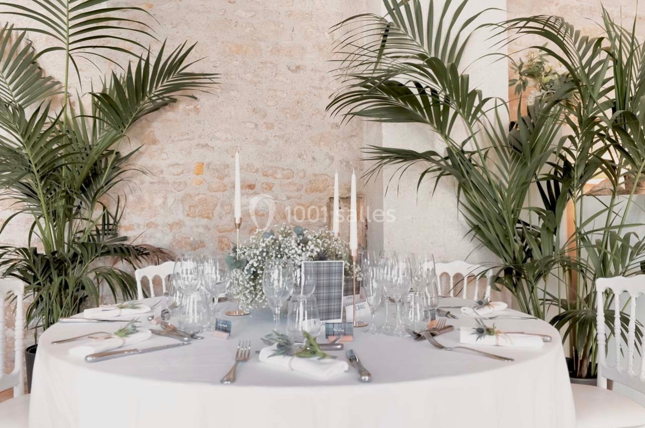 Table élégamment dressée avec nappes blanches, couverts raffinés, bougies et décorations florales, entourée de plantes…
