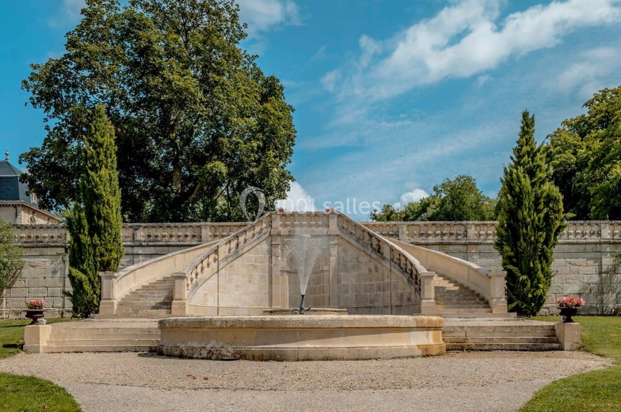 Escalier en pierre symétrique entouré d'arbres, avec une fontaine centrale et un ciel bleu en arrière-plan.