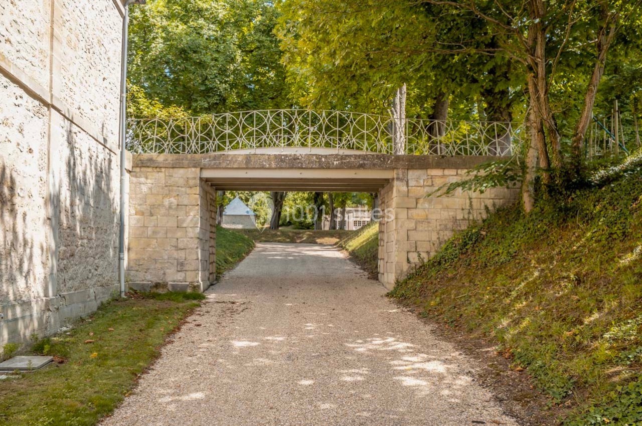 Petit pont en pierre avec balustrade métallique, surplombant un chemin bordé d'herbe et d'arbres.
