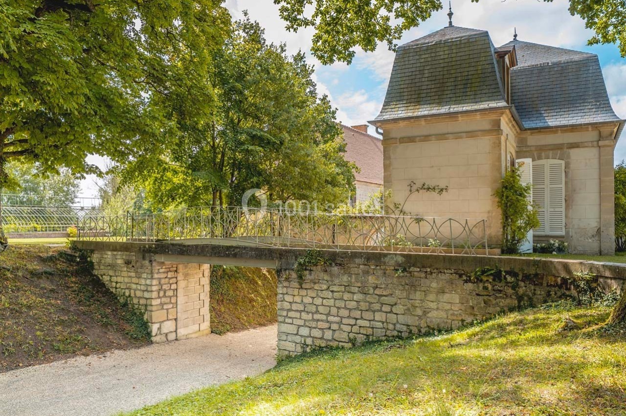 Petit pont en pierre surplombant un chemin, entouré de verdure et jouxtant un bâtiment ancien avec toiture en ardoise.