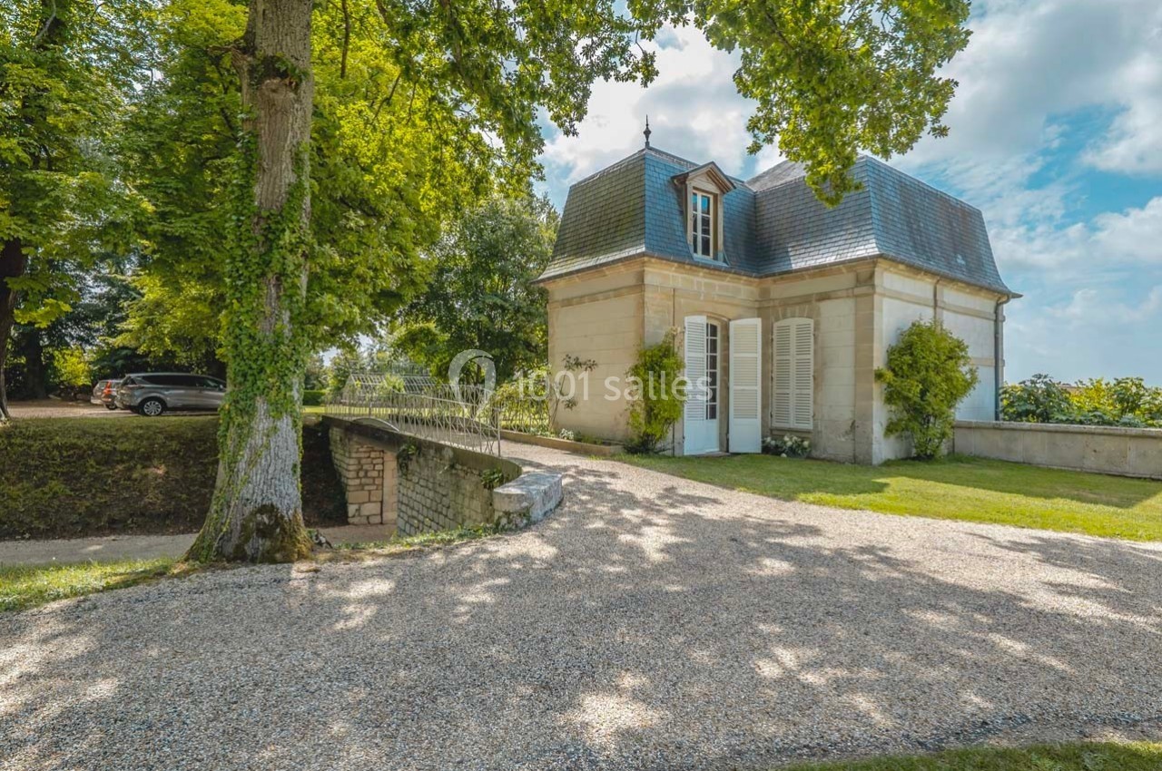 Petite maison en pierre avec volets blancs, entourée de verdure, près d'un chemin gravillonné et d'un pont en pierre.