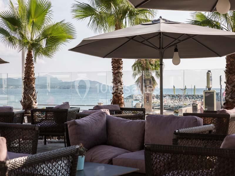 Terrasse avec fauteuils en osier, parasols et vue sur la mer, entourée de palmiers et d'une barrière en verre.