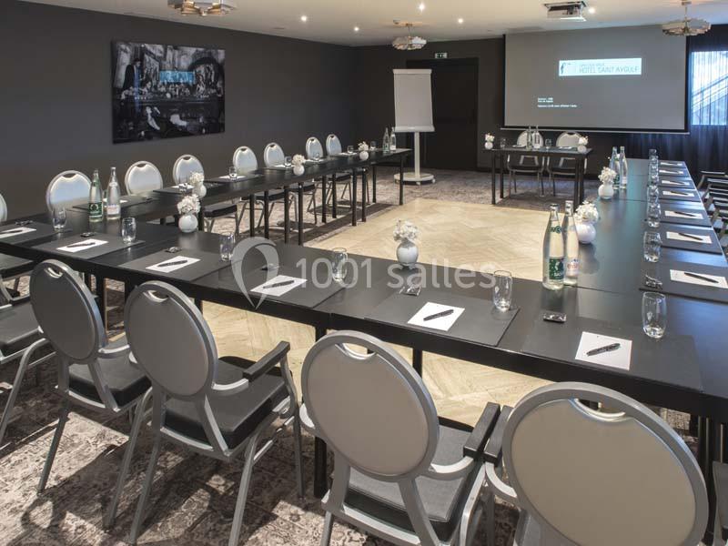Salle de réunion moderne avec tables disposées en U, chaises grises, écran de projection et matériel de conférence.