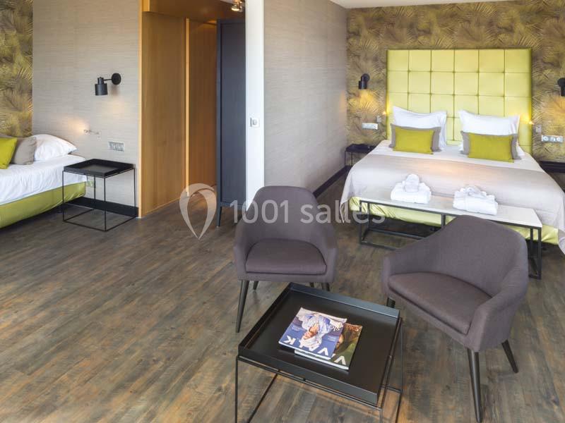 Chambre d'hôtel moderne avec un lit double, un lit simple, deux fauteuils et une table basse sur un sol en parquet.