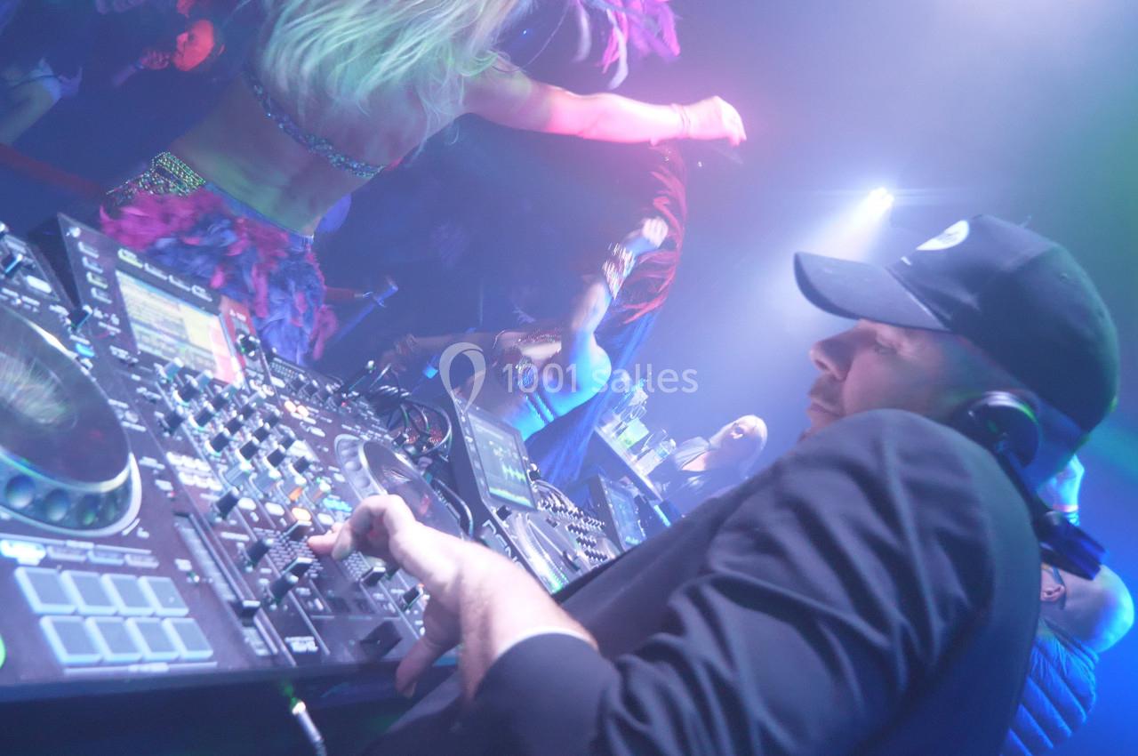 Un DJ mixe sur une console dans une ambiance de fête avec des danseurs et des lumières colorées en arrière-plan.