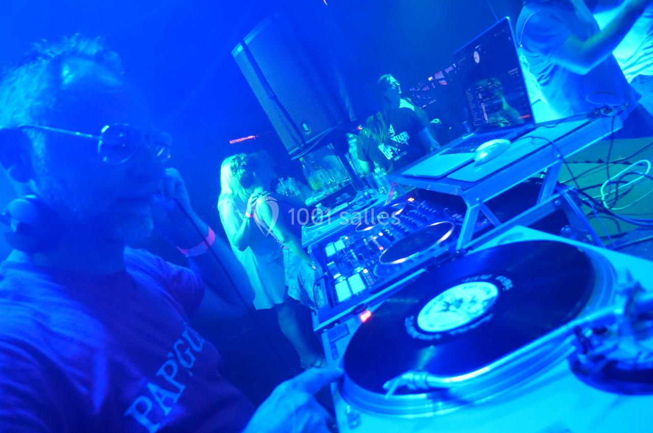 Un DJ mixe des vinyles sur une platine dans une ambiance lumineuse bleutée, entouré de personnes dansant.