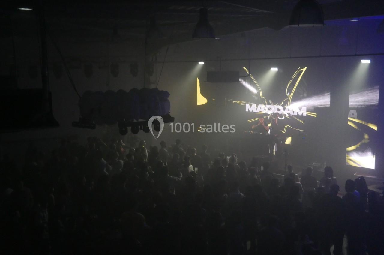 Salle sombre avec une foule devant une scène éclairée par des projections lumineuses et un écran affichant ’MADDAM’.