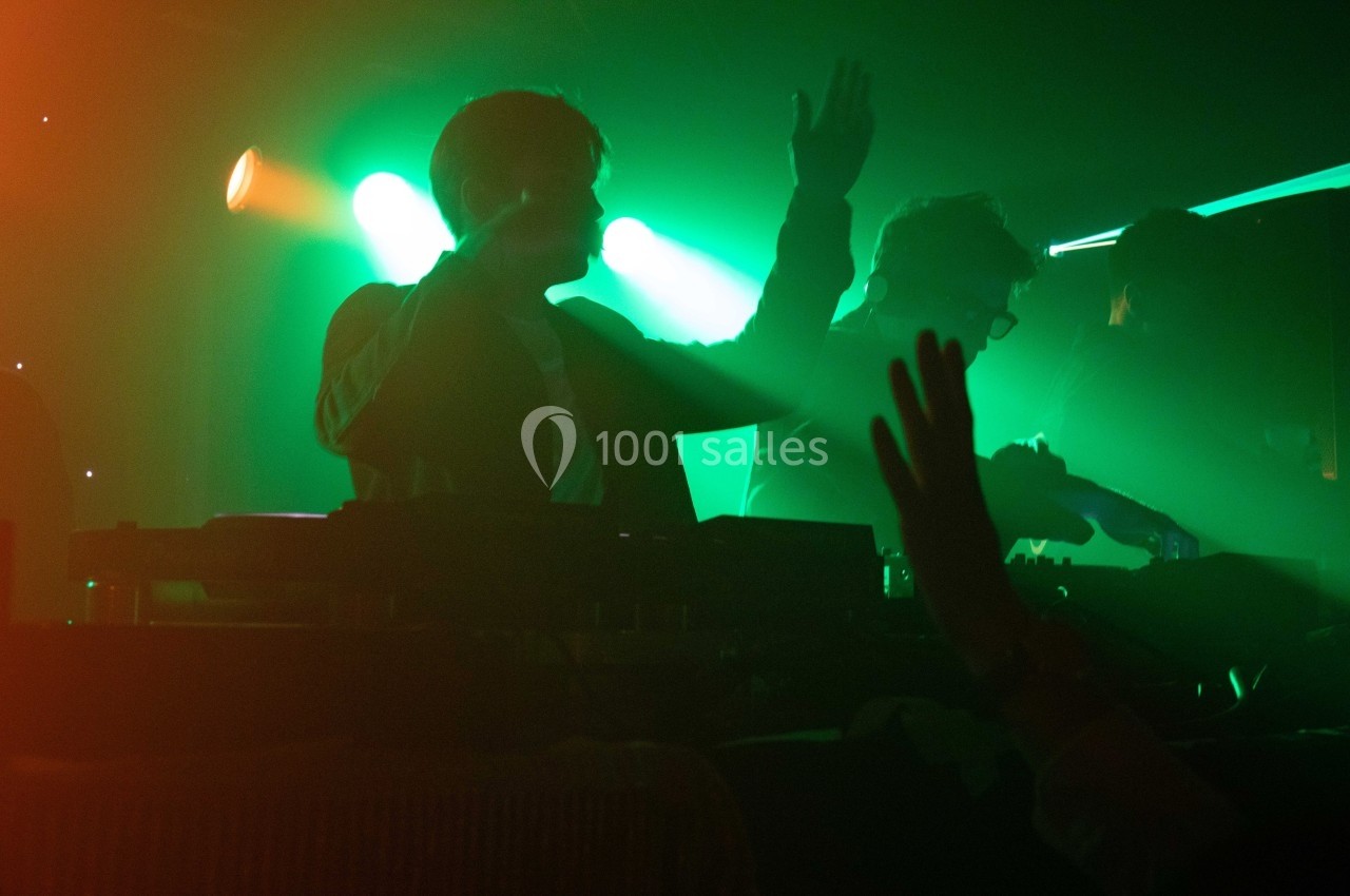 Un DJ mixe sur scène dans une ambiance lumineuse verte et orange, entouré d'un public avec des mains levées.