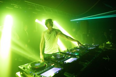 Miniature Equinoxe #27 Un DJ mixe sur une scène éclairée, avec un écran lumineux affichant des motifs rouges en arrière-plan.