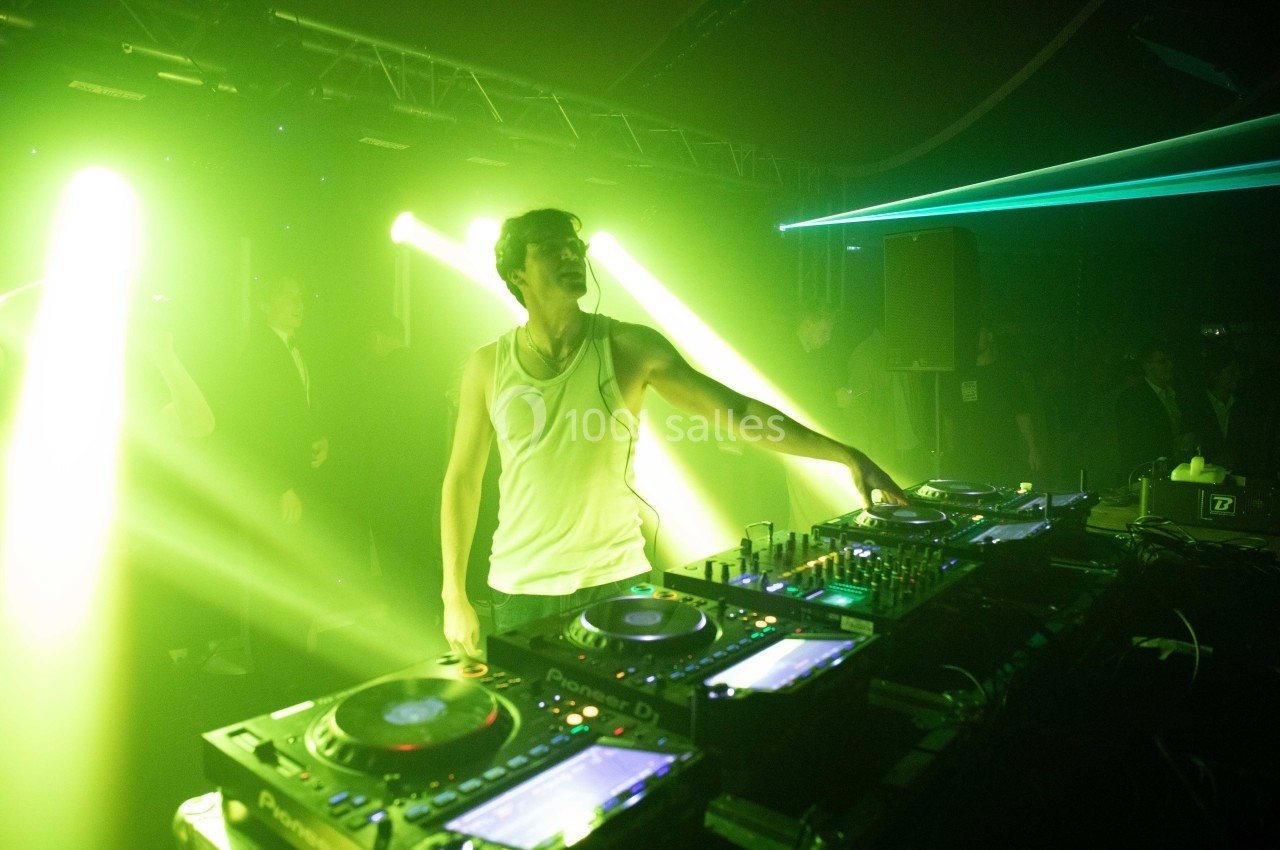 Un DJ en débardeur blanc mixe sur une table de mixage dans une ambiance lumineuse verte et sombre.