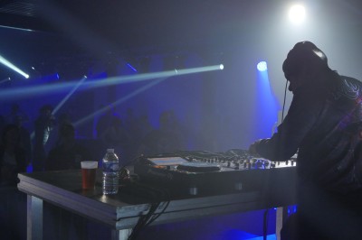 Miniature Equinoxe #37 Un DJ mixe sur une scène éclairée, avec un écran lumineux affichant des motifs rouges en arrière-plan.