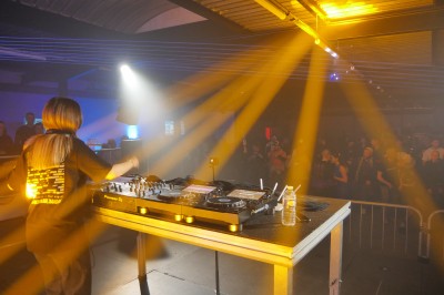 Miniature Equinoxe #38 Un DJ mixe sur une scène éclairée, avec un écran lumineux affichant des motifs rouges en arrière-plan.